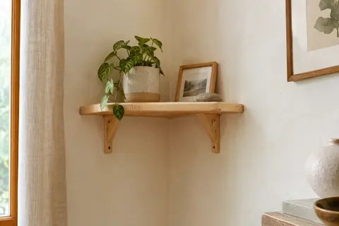 Petite étagère d’angle en pin naturel fixée sur un mur blanc cassé, avec supports et objets décoratifs.